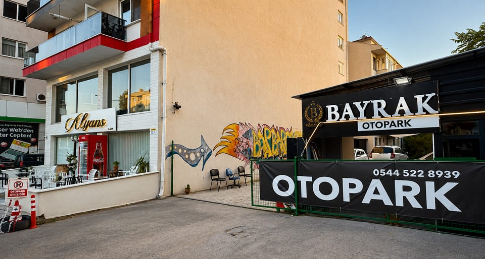 Bayrak Otopark'ın Atatürk Caddesi üzerindeki girişi: aydınlatmalı tabela, bariyerli kapı ve çevre çiti